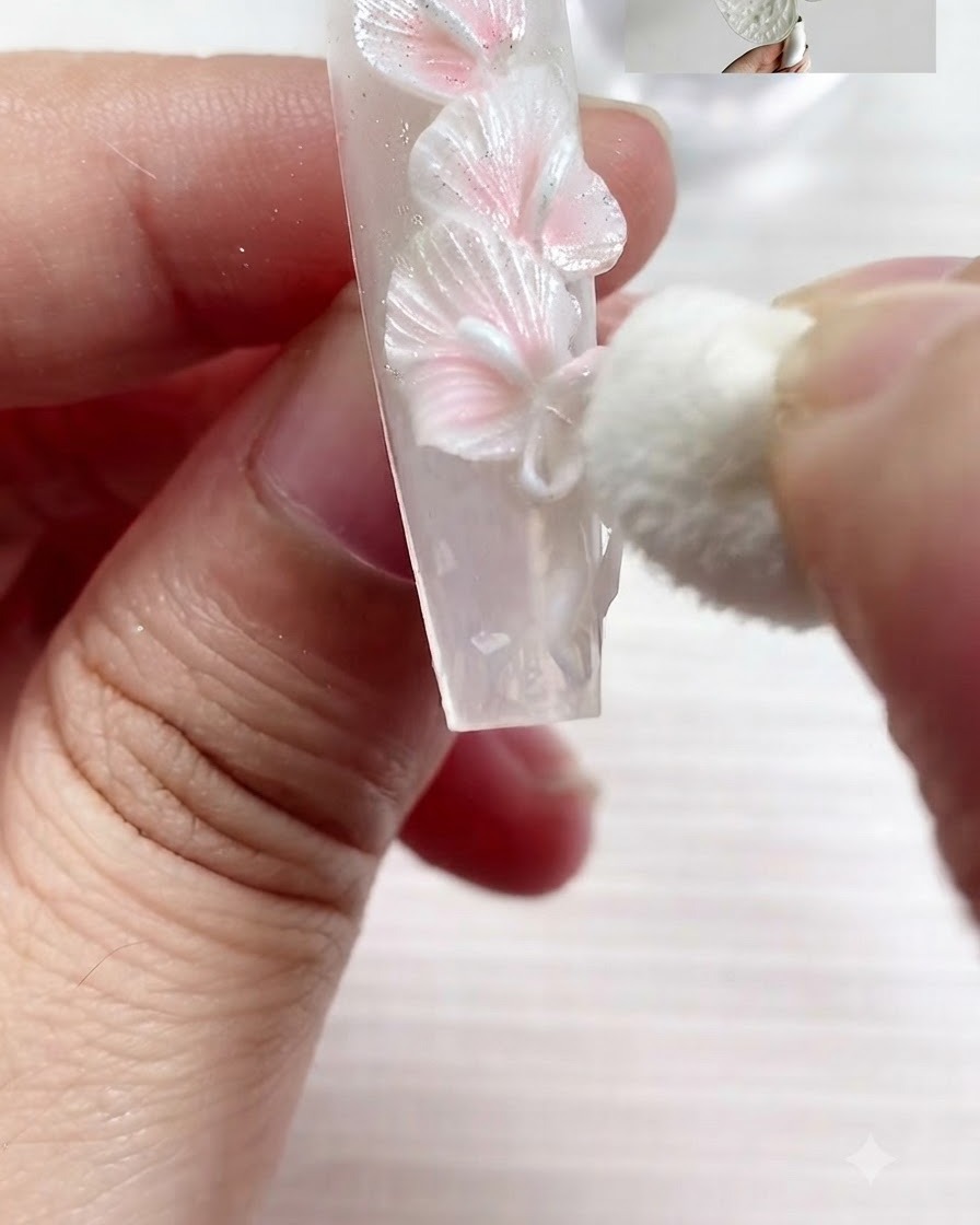 Mała gąbka kosmetyczna delikatnie wklepująca perłowo-biały pigment na teksturowane krawędzie wyrzeźbionych białych kalii i kokardki na paznokciu.