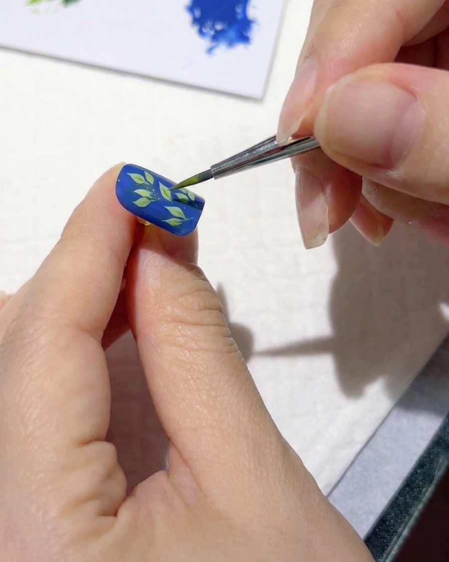 Cienki pędzelek do nail artu dodający jasnozielone pociągnięcia nad ciemnymi liśćmi na niebieskim paznokciu.