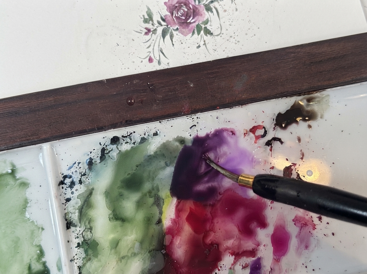 Cienki pędzelek do akwareli mieszający ciemnofioletowy pigment na czystej białej palecie obok zielonych lawunków.