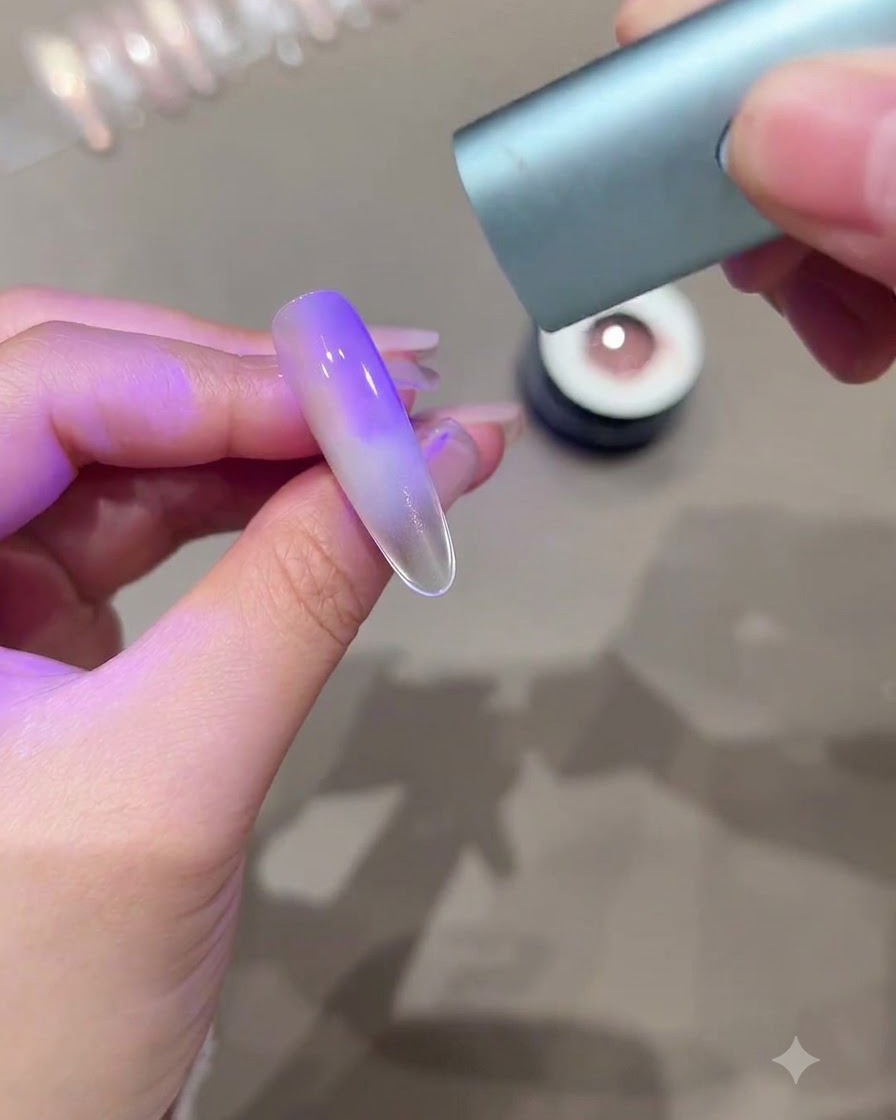 Ruka držiaca mini UV LED vytvrdzovacie svetlo nad predĺženým nechtom zobrazujúcim fialový gradientný efekt mačacieho oka.