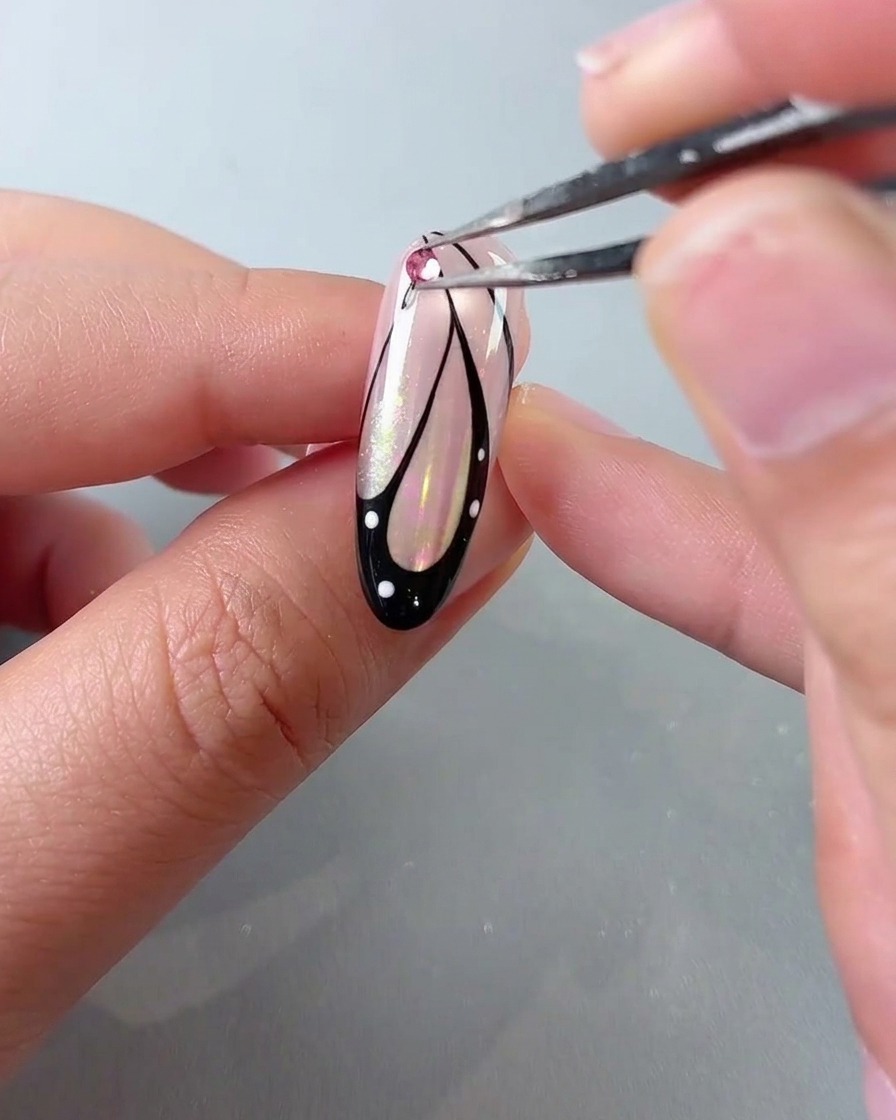 Eine Pinzette platziert einen rosa Kristall-Strassstein oben auf einem Schmetterlingsflügel-Nail-Art-Design.