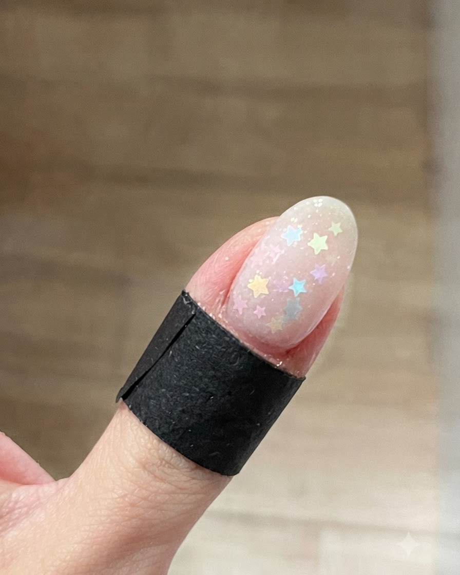 Eine Nahaufnahme eines ovalen Fingernagels mit mehrfarbigen Pastell-Stern-Glitzern, die auf einem klaren, feuchten Base-Gel verstreut sind.