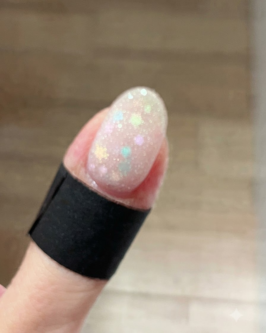 Eine Nahaufnahme eines Fingernagels, der mit einem feuchten, funkelnden frostweißen Schimmergel über Pastell-Stern-Glitzern beschichtet ist.