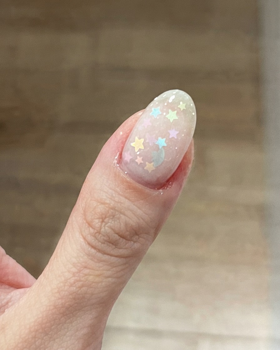 Eine Nahaufnahme eines Daumennagels mit einer milchig-roten Basis und darauf platzierten bunten Pastell-Stern-Glitzern.