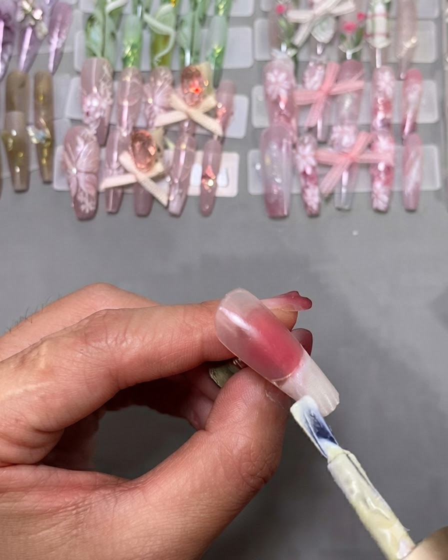 Standardowy pędzelek do lakieru hybrydowego aplikujący biały lakier na końcówkę sztucznego paznokcia zamontowanego na stojaku.