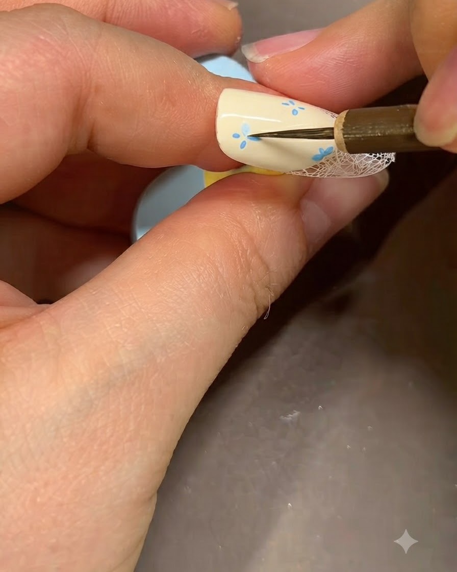 Cienki pędzelek do nail art dodający szczegółowe niebieskie akcenty w środku namalowanego kwiatu na beżowym paznokciu.