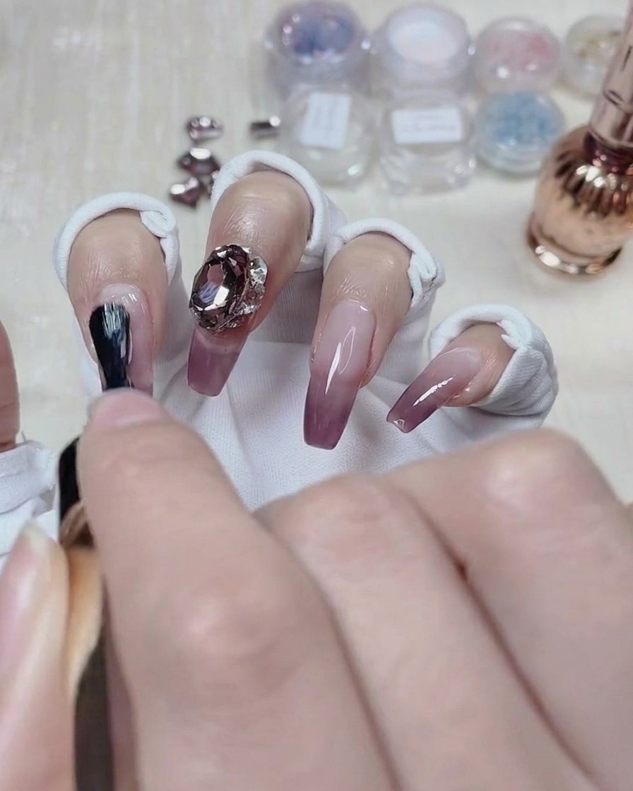 Stylista paznokci nakładający top coat wokół podstawy kryształowej ozdoby na wykończonym paznokciu z gradientem.