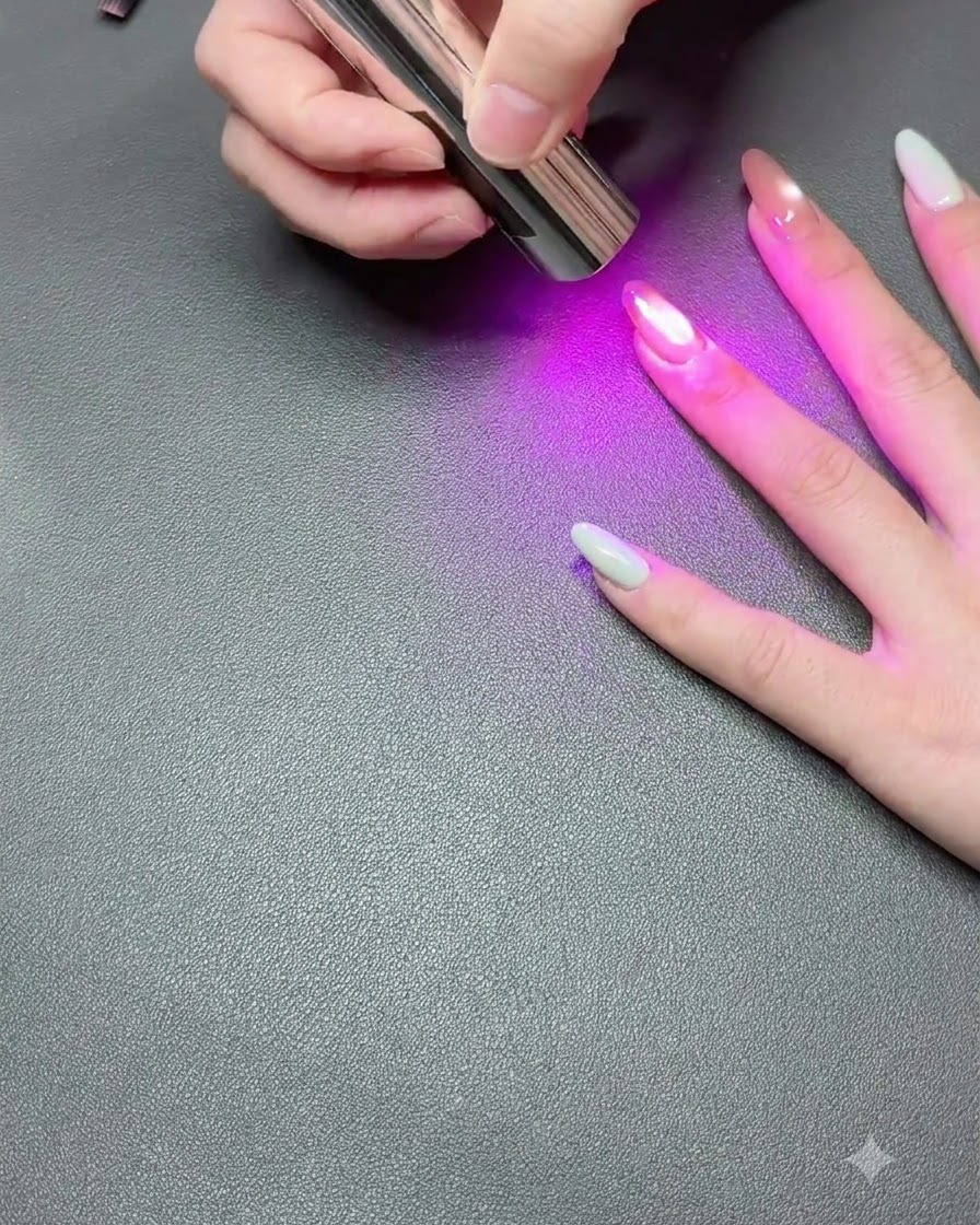 Dłoń spoczywająca na ciemnej powierzchni, podczas gdy przenośna lampa UV utwardza lakier hybrydowy cat-eye.