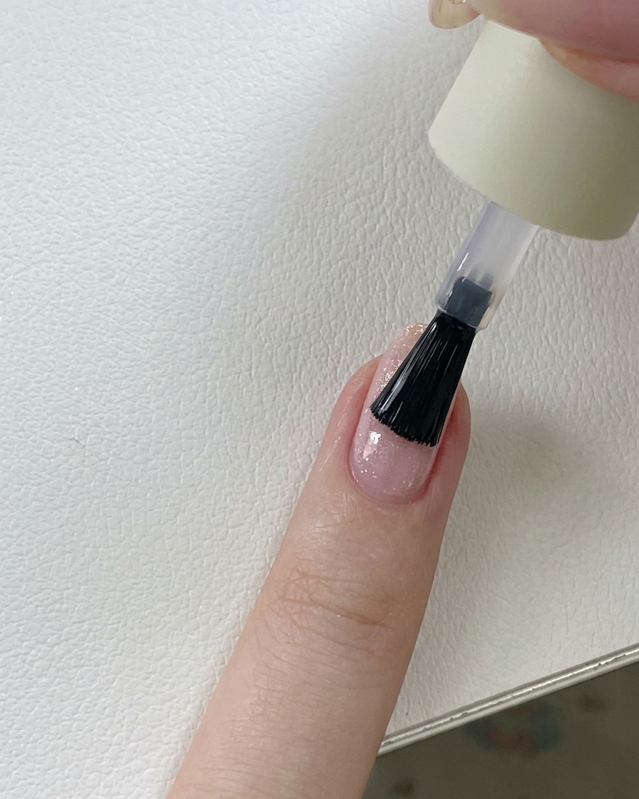 Pędzelek nakładający klarowny, błyszczący top coat na różowy paznokieć z brokatem żelowym.