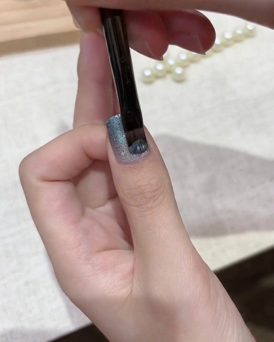 Ein Gelpinsel trägt eine Schicht schimmernden silber-blauen magnetischen Cat-Eye-Lacks über eine Gradienten-Basis auf.
