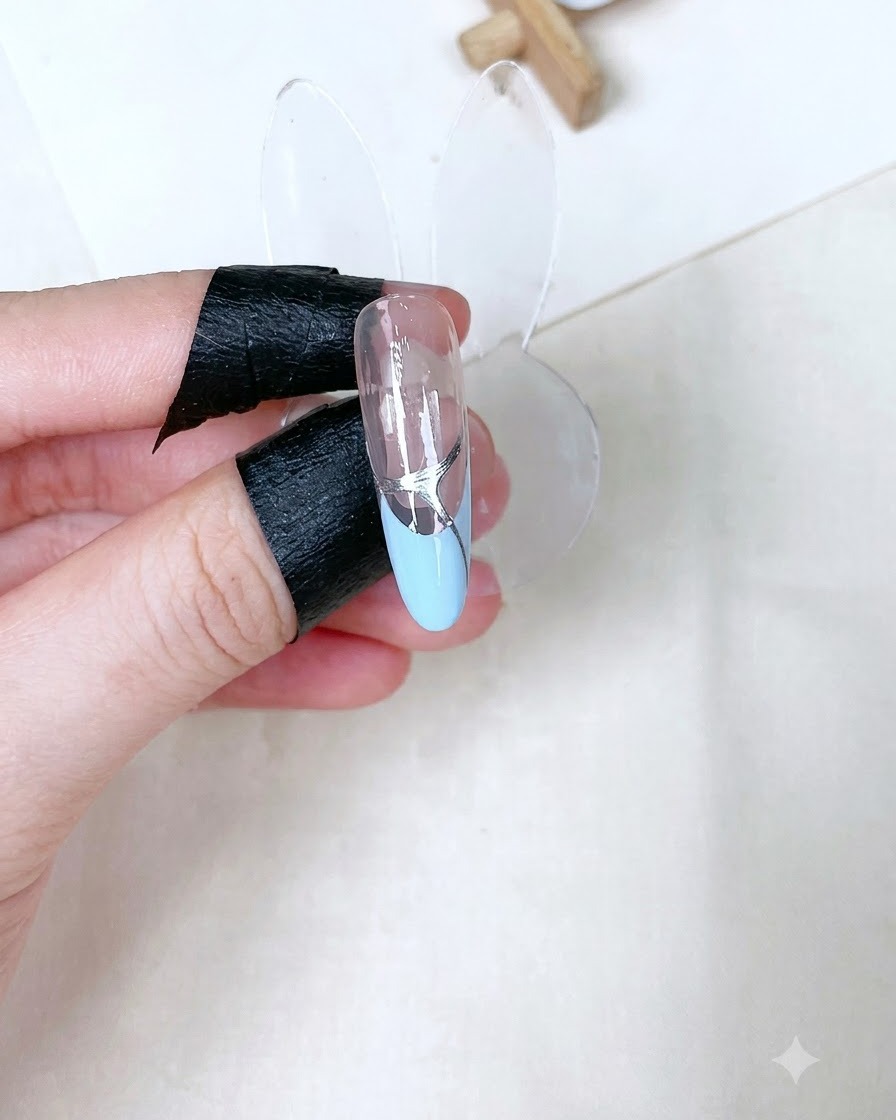 Pojedynczy wyrzeźbiony stojak na paznokcie ukazujący utwardzony, błyszczący, przezroczysty top coat ze srebrnymi detalami nad frenchem baby blue.