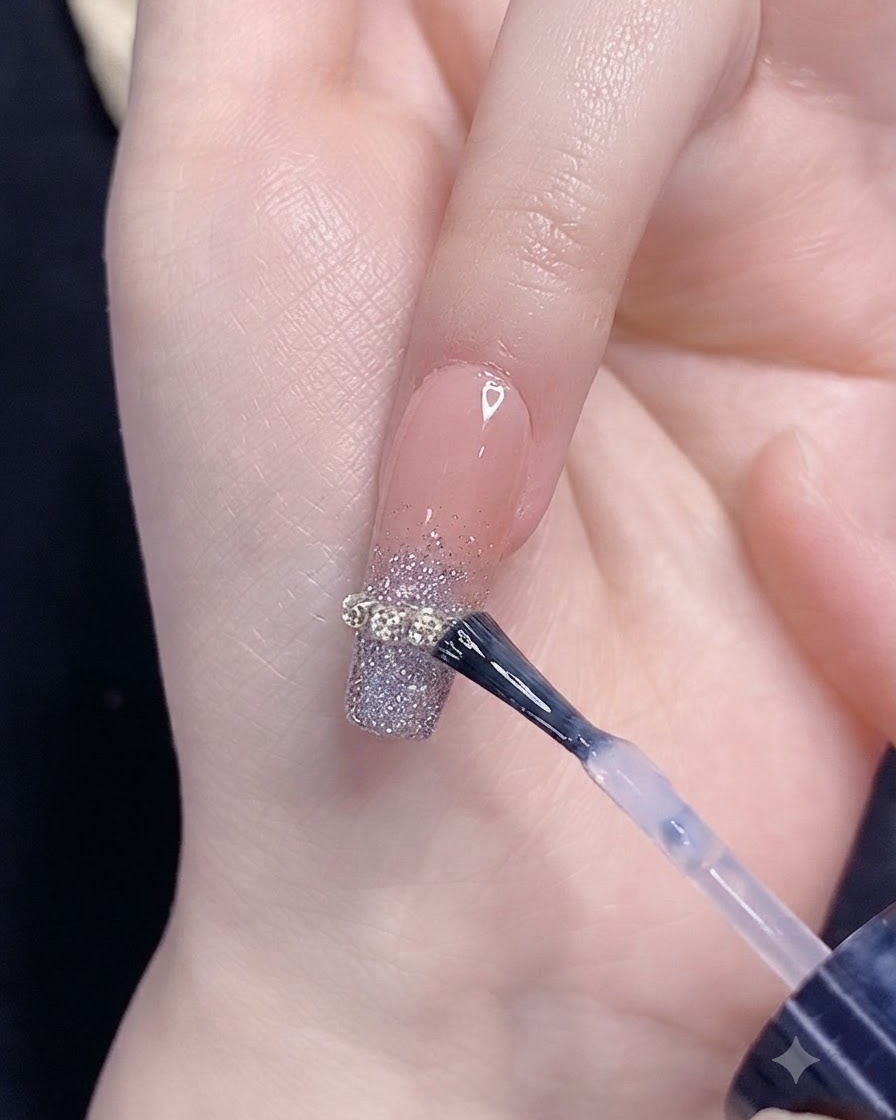 Pędzelek nakładający top coat na paznokieć ze srebrnym brokatowym gradientem i ozdobą z łańcuszka.