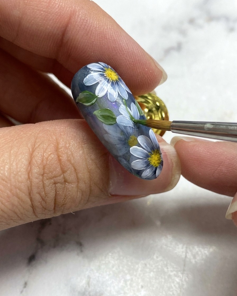 Pędzelek do nail art dodający mały zielony liść obok białych detali kwiatowych na szarej bazie paznokcia.