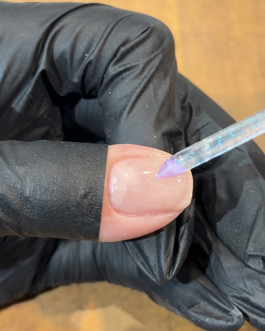 Dłoń w rękawiczce używająca cienkiego pędzelka do umieszczenia kropli niebieskiego żelu obok fioletowej sekcji na paznokciu.