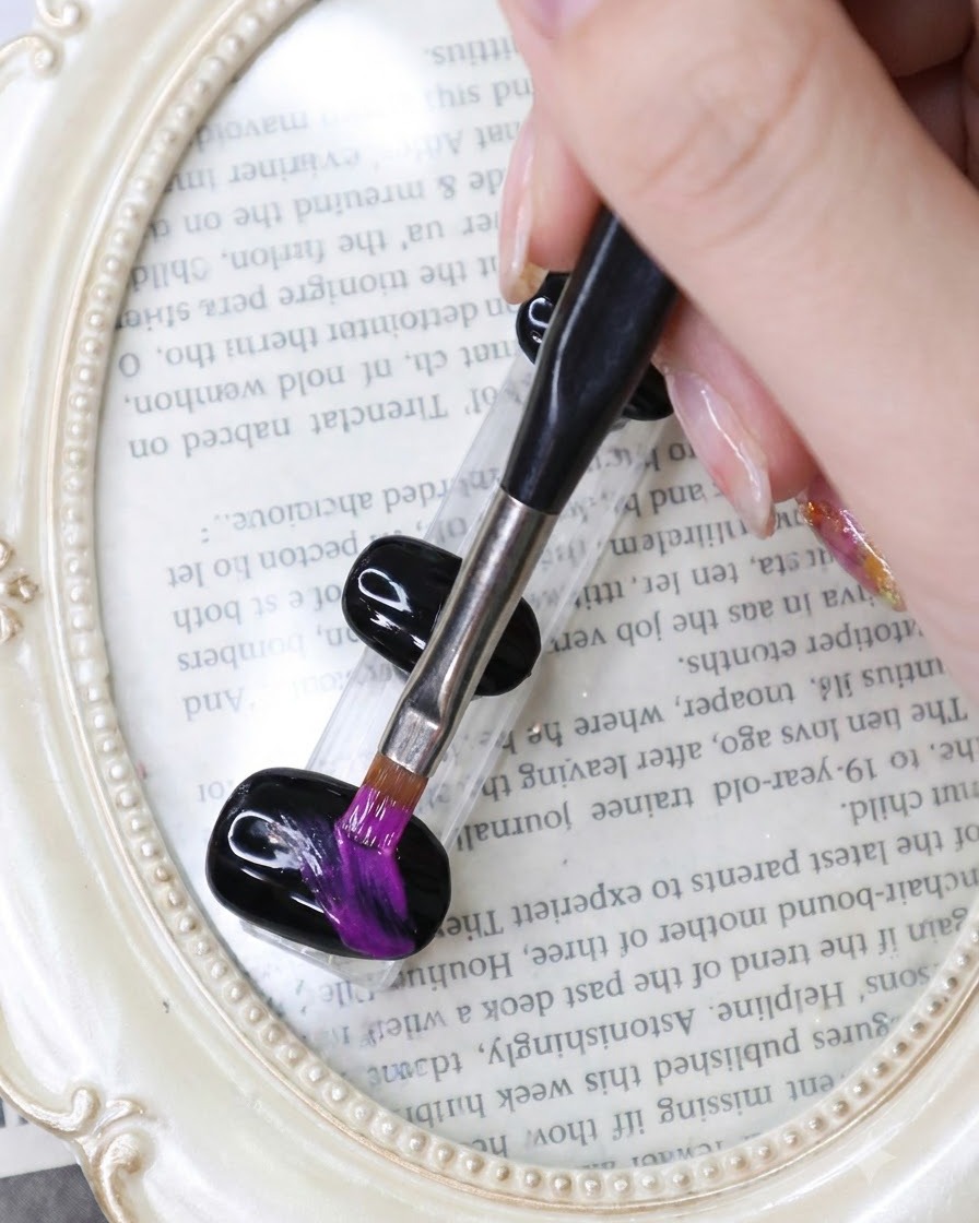 Ein feiner Liner-Pinsel zieht lebendige Neon-Lila-Pinselstriche über einen schwarzen Gel-Nagel vor einem buchbezogenen Hintergrund.