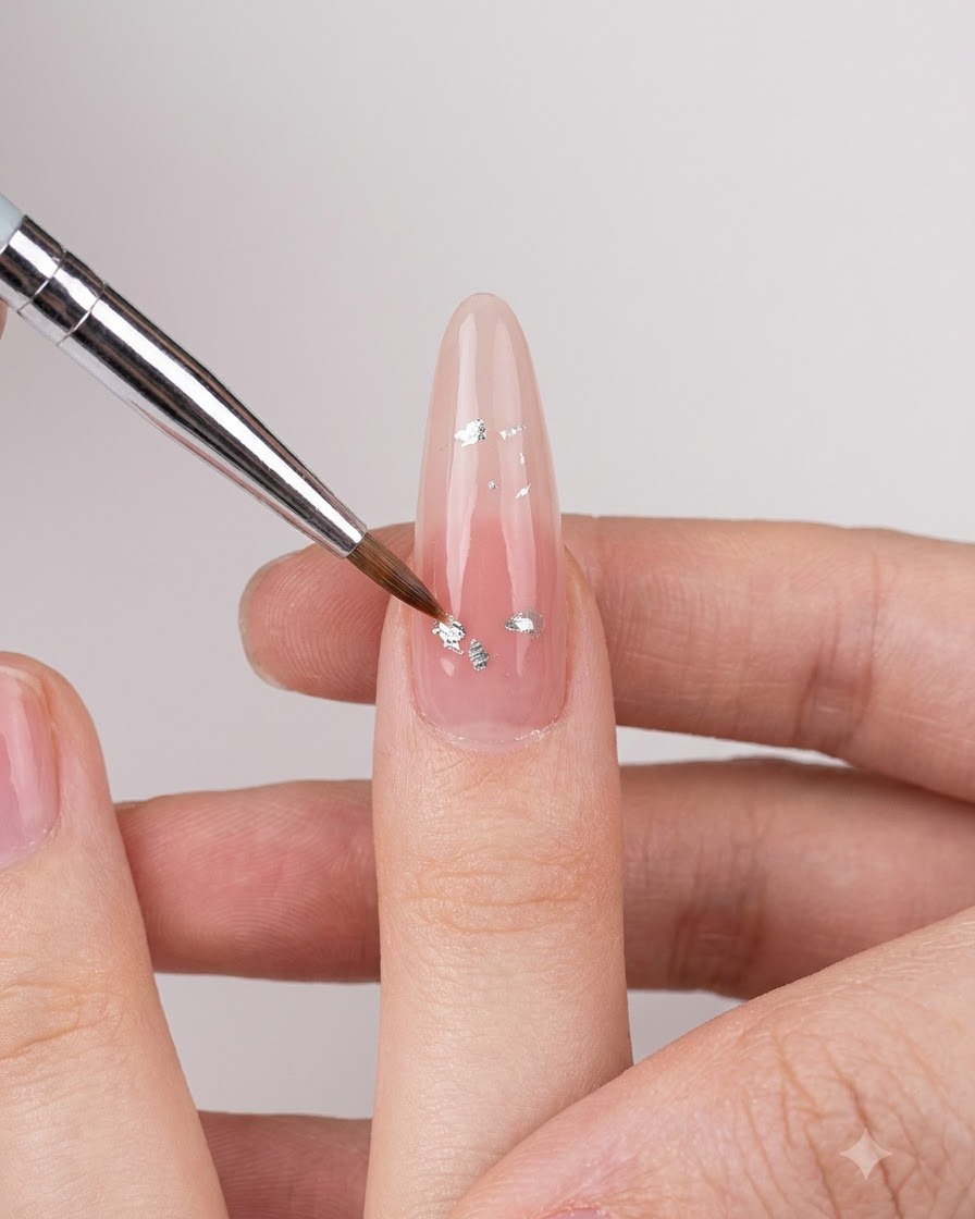 Cienki pędzelek do nail art nakładający małe srebrne iryzujące płatki na cielisty błyszczący paznokieć.