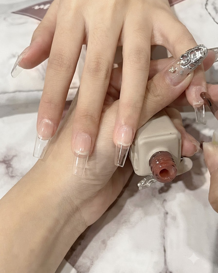 Pędzelek do paznokci nakładający cienką warstwę przezroczystego żelu nude na długą, naturalną płytkę paznokcia o kształcie trumienki.