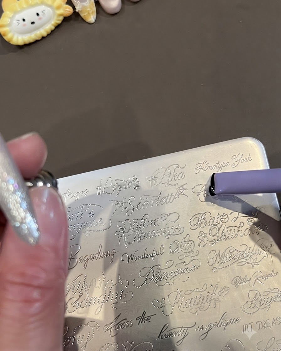 Zbliżenie na metalową płytkę do stempli z wygrawerowanymi kursywą angielskimi słowami oraz fioletowy żelowy stamper zbierający wzór.