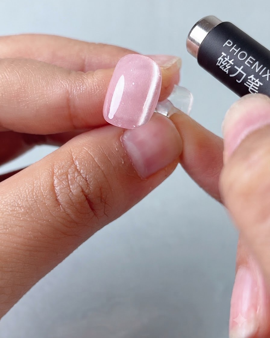 Narzędzie magnetyczne w formie długopisu unoszące się nad różowym paznokciem żelowym cat-eye w celu uformowania pasa świetlnego.