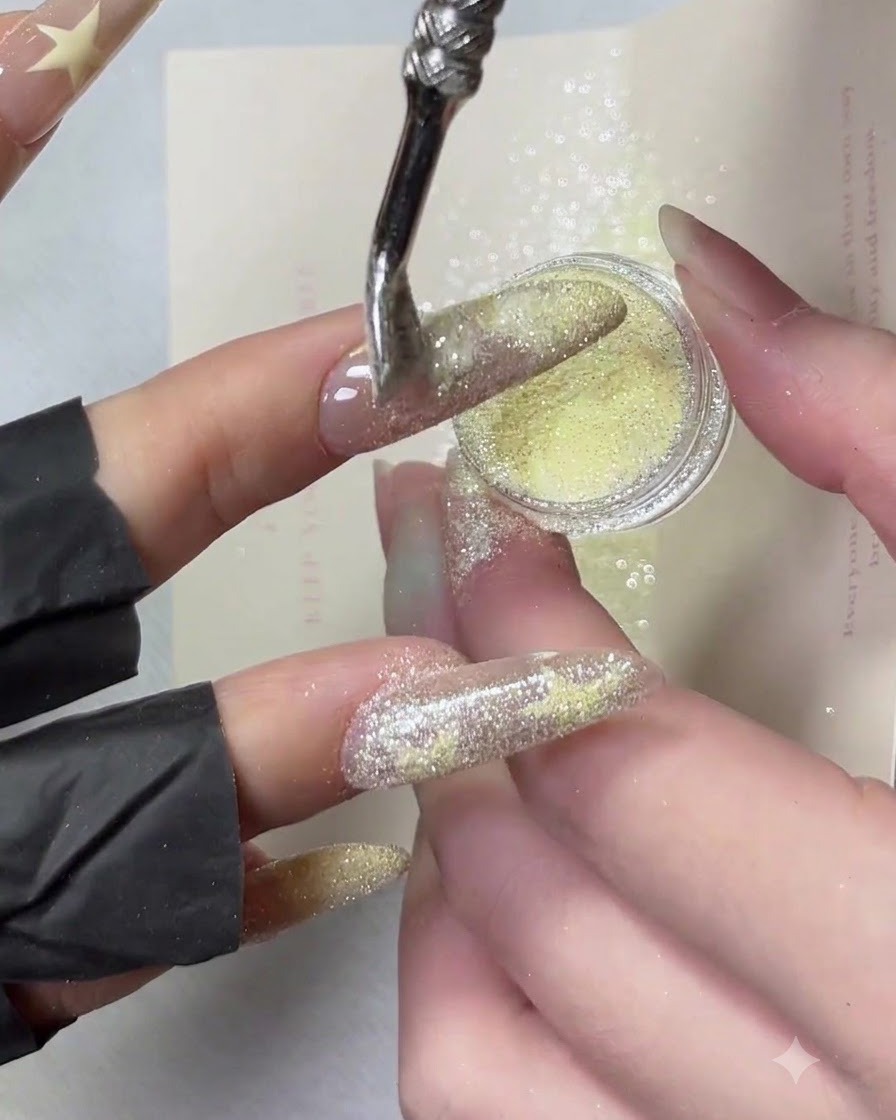 Dłoń przechylona bokiem, podczas gdy żółty pyłek crushed diamond jest oprószany wzdłuż krawędzi końcówki manicure french.